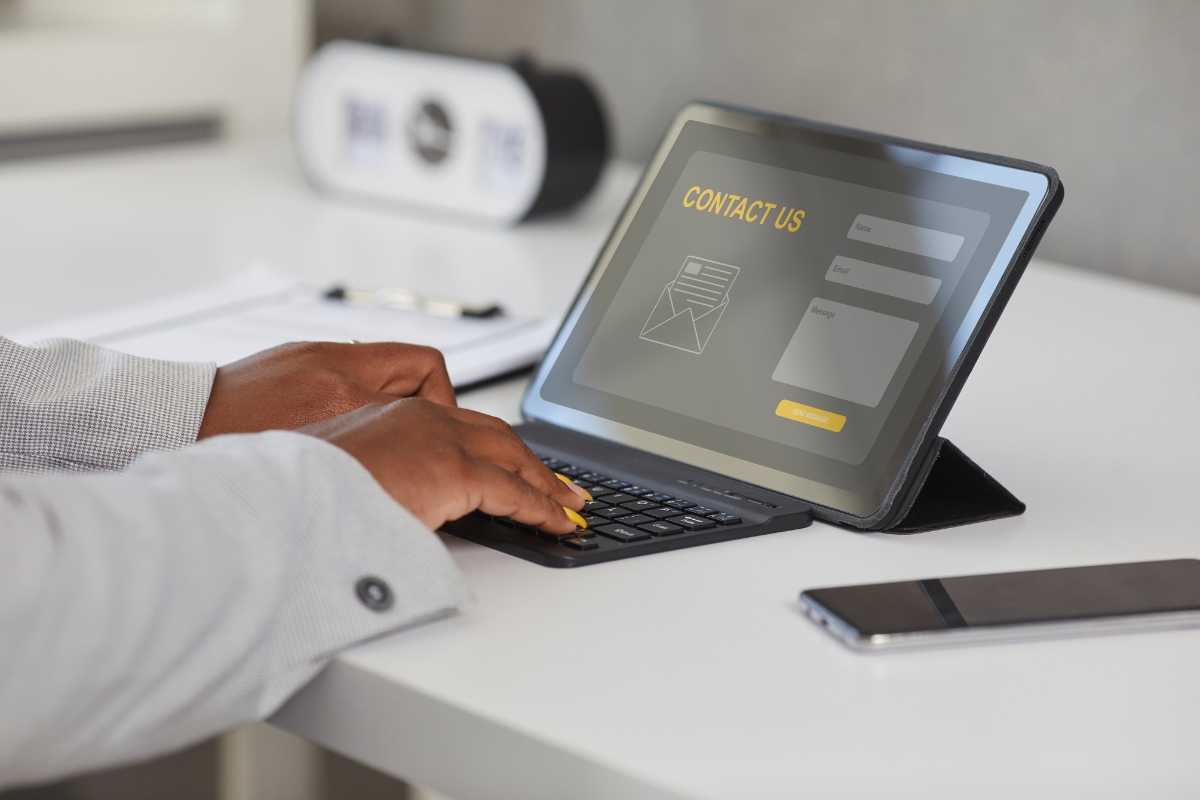 A person types on a tablet's keyboard at a desk. The tablet screen displays a "Contact Us" form, much like what you would find on a landing page, and a smartphone lies nearby.