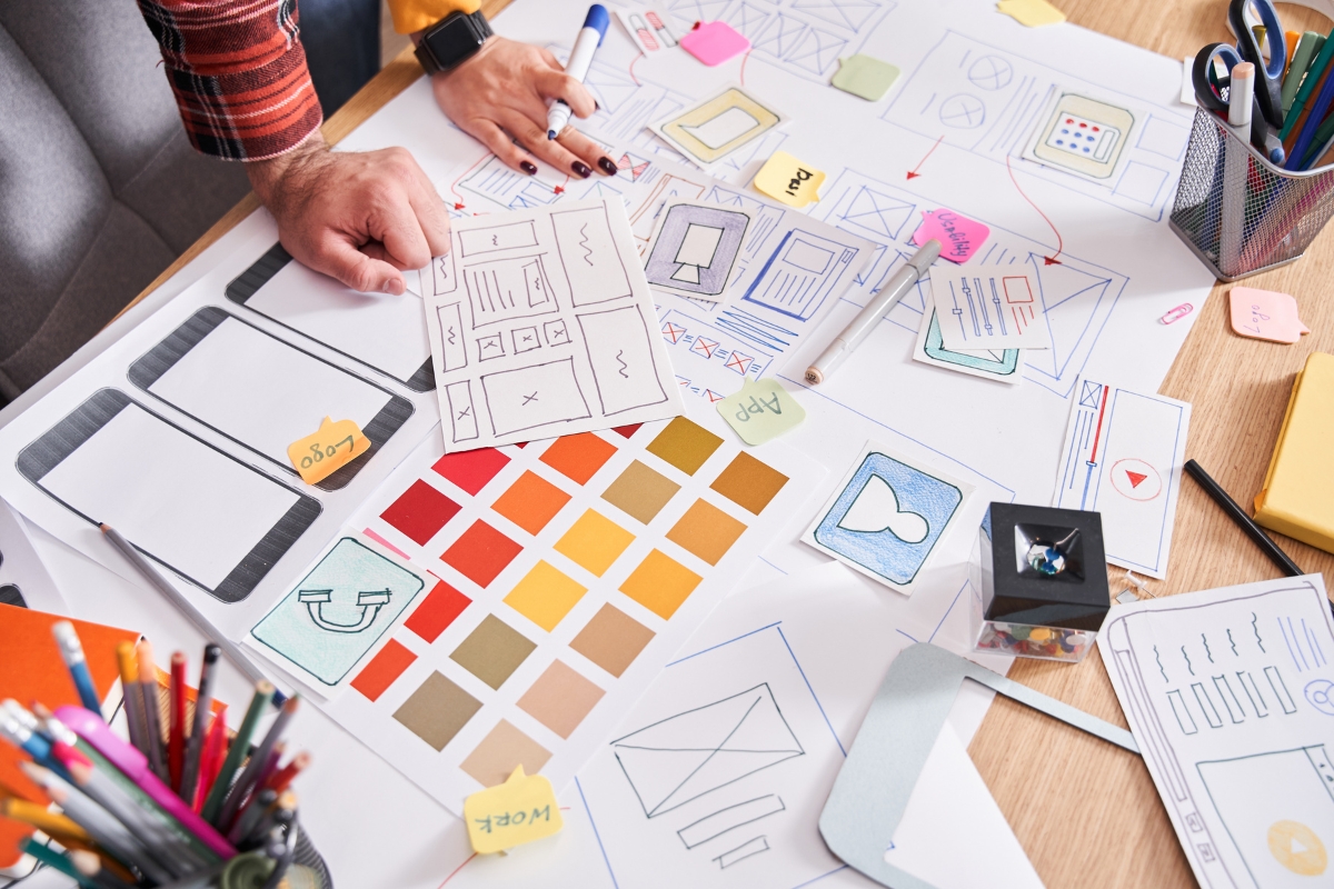 People working on a website design project with various papers, sketches, color swatches, and sticky notes on a cluttered desk are likely discussing what is a landing page to ensure its effectiveness in guiding visitors.