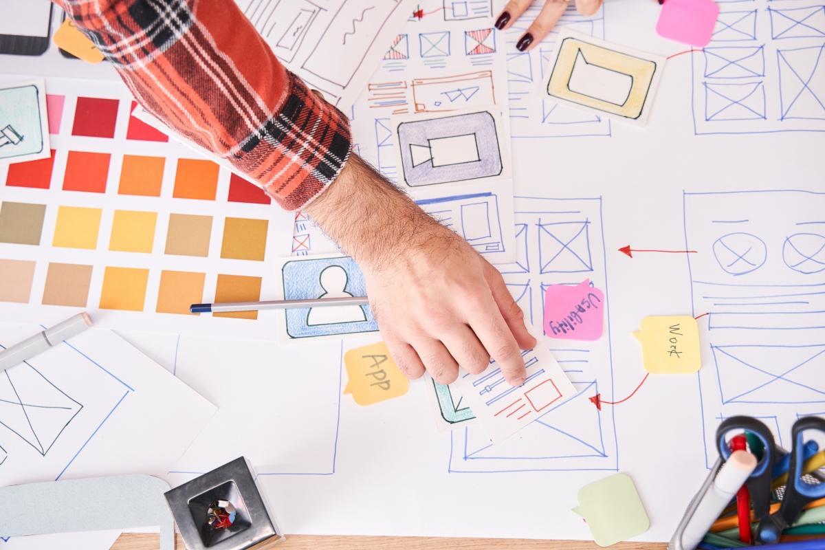 A person in a red plaid shirt is arranging paper prototypes for an app's user interface on a cluttered desk covered with design sketches, sticky notes, and color swatches, pondering *what is a landing page* to improve the design.