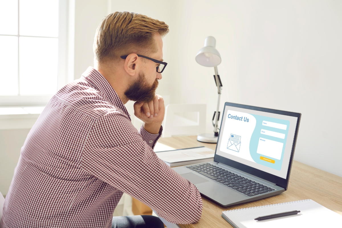 A man with glasses and a beard is sitting at a desk, looking at a laptop screen displaying a "Contact Us" form, wondering what is a landing page.