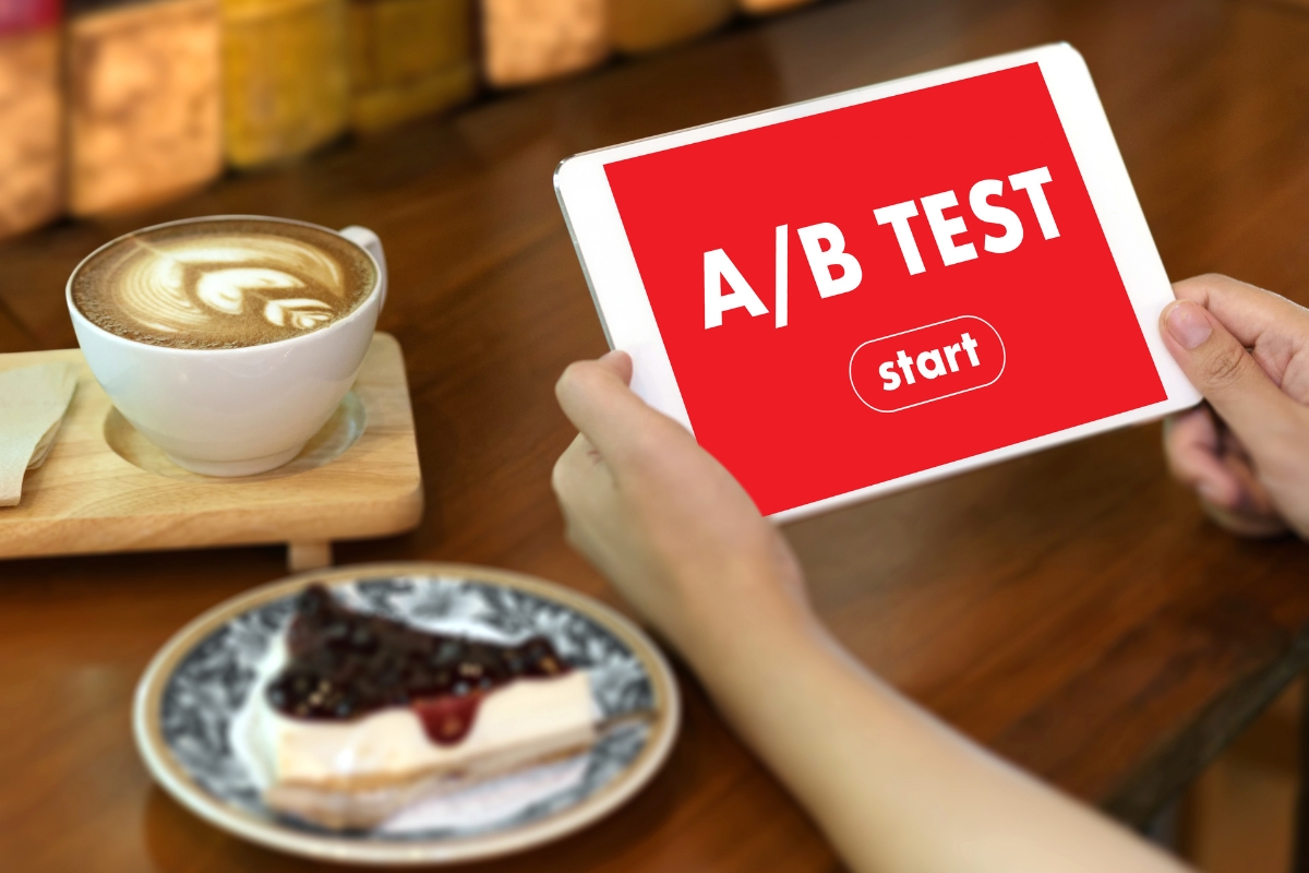 A person holding a tablet displaying "A/B TEST" with a start button. A cup of latte and a slice of cheesecake are on the table, making one wonder, "What is AB testing in marketing?