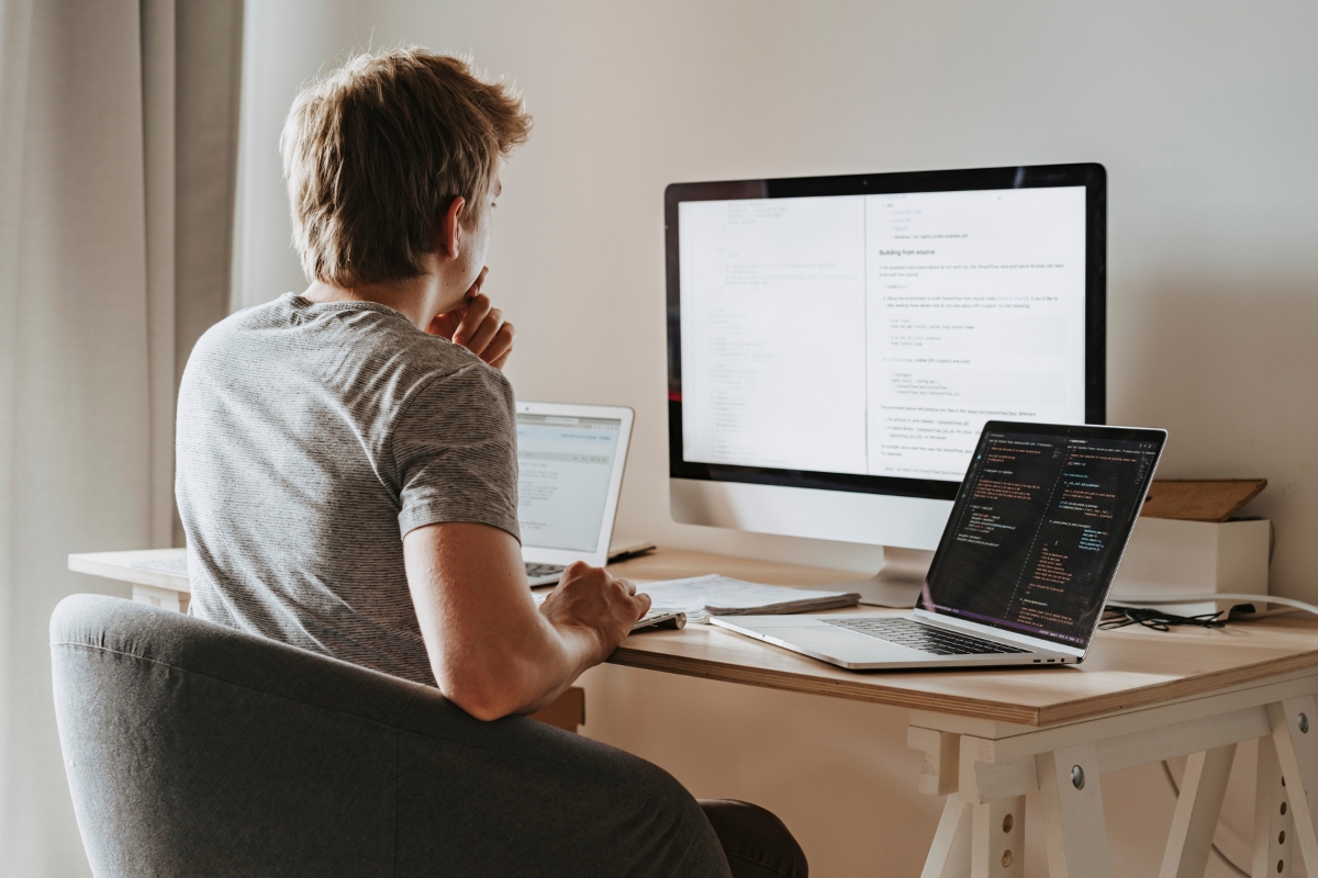 A person is seated at a desk working on three computer screens. The screens display code and documents, possibly related to questions like "what is AB testing in marketing?" The setting appears to be a home or office workspace.