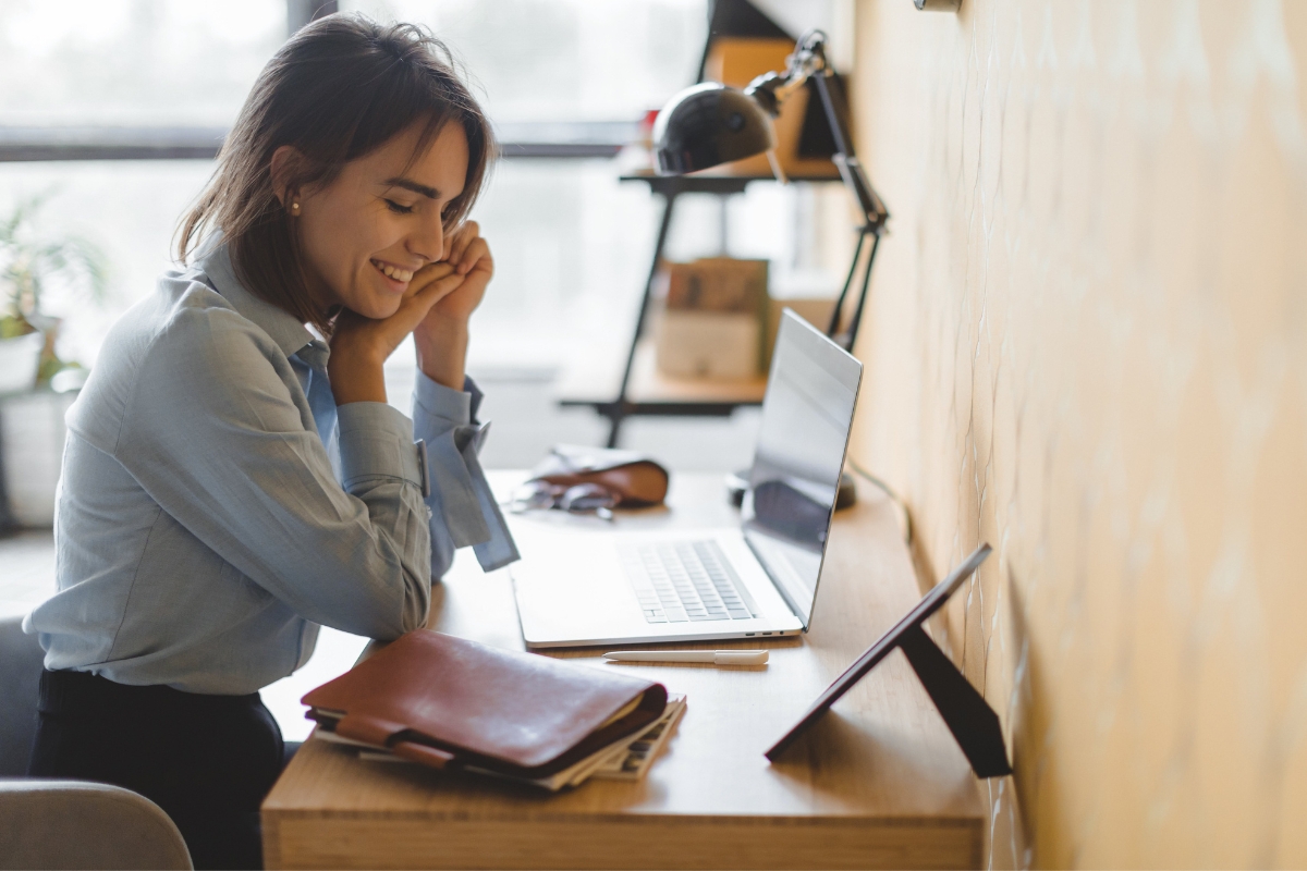 A woman sits at a desk, smiling while looking at a laptop screen, with a tablet, notebook, and glasses on the desk. She seems engrossed in her research on "what is AB testing in marketing.