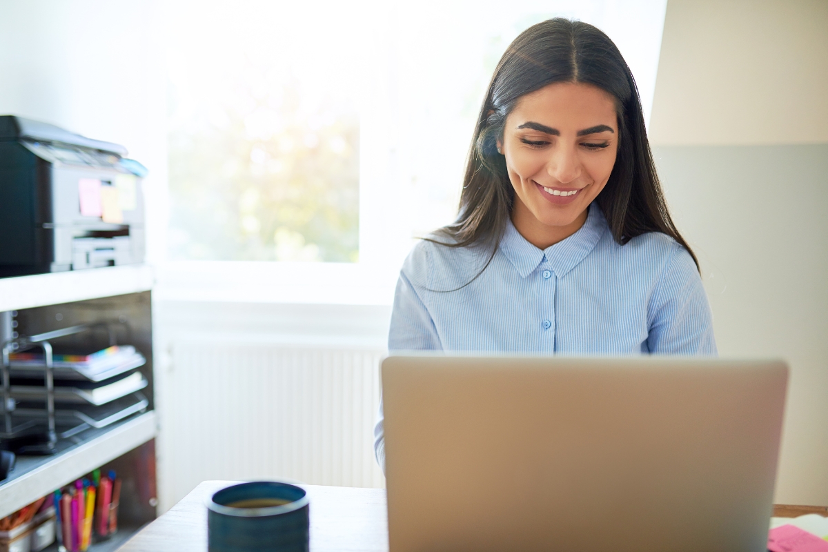 A woman sits at a desk with a smile, working on a laptop in a bright, sunlit room. A cup and printer are visible in the background as she possibly researches what is AB testing in marketing.