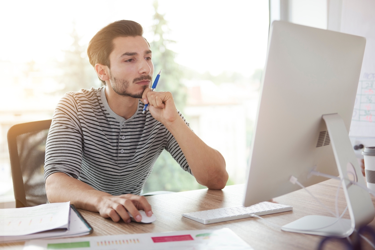 A man wearing a striped shirt sits at a desk, looking at a computer screen with a pen in his hand. He appears to be concentrating on his work, possibly researching what is A/B testing.