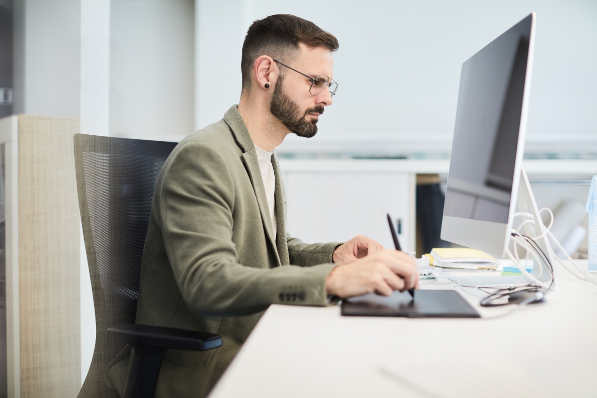 Man with glasses and beard working on a computer at a desk in a modern office environment. He is using a stylus and graphic tablet, seemingly focused on designing visuals for an upcoming project. Occasionally, he glances at another screen displaying information about what is A/B testing in marketing.