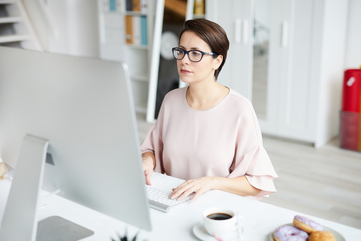 A woman with glasses is seated at a desk, working on a computer. There is a cup of coffee and some doughnuts on the desk. She appears to be focused on the screen, perhaps researching what AB testing in marketing entails.