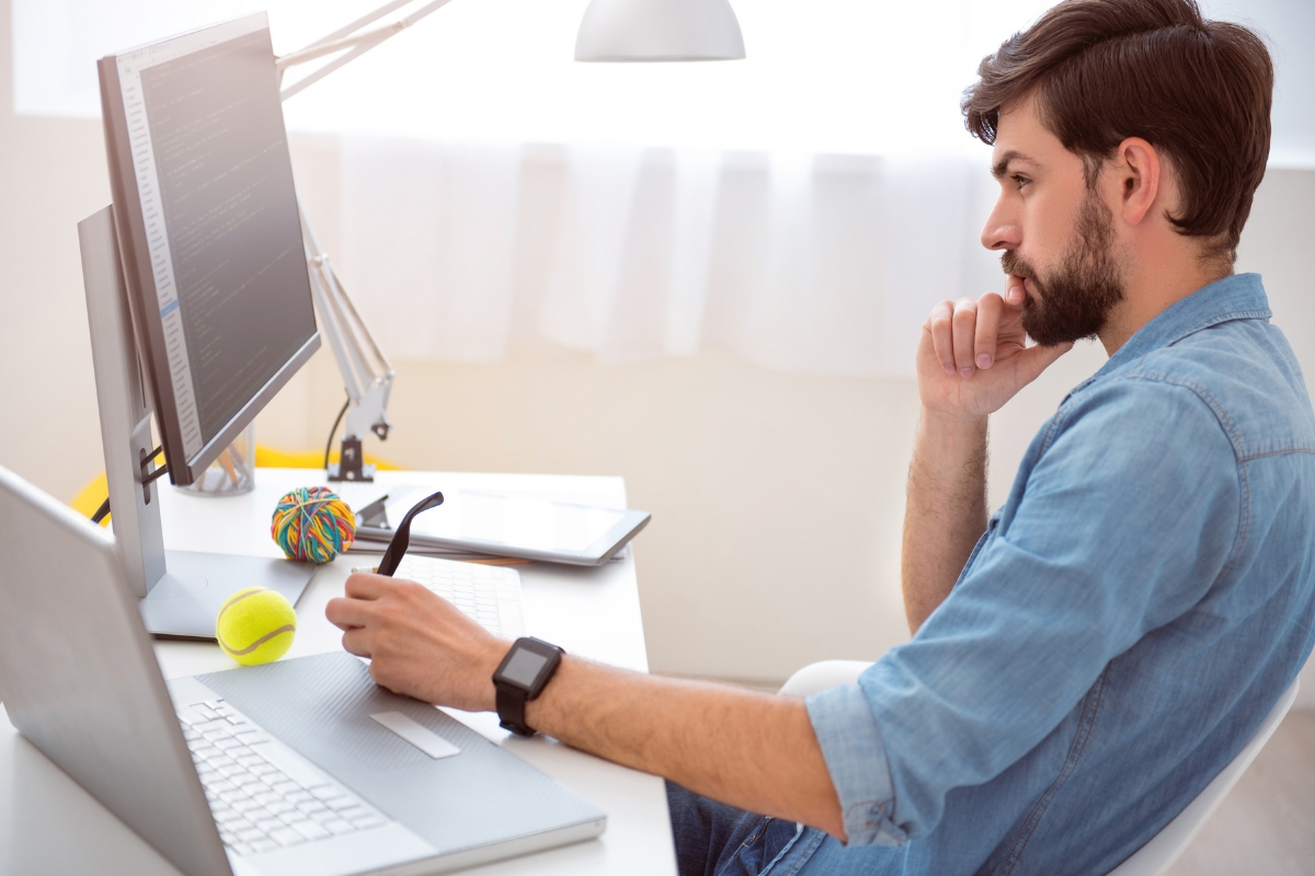 A man wearing a denim shirt and smartwatch is sitting at a desk with multiple computer monitors, focused on coding. Nearby, a tennis ball and a colorful stress ball lie on the desk, perhaps to help him think about what is AB testing in marketing for his next project.