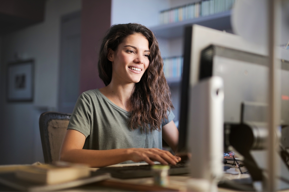 A woman with long hair and a green shirt is smiling while using a desktop computer, possibly researching what A/B testing in marketing is. Bookshelves are visible in the background.