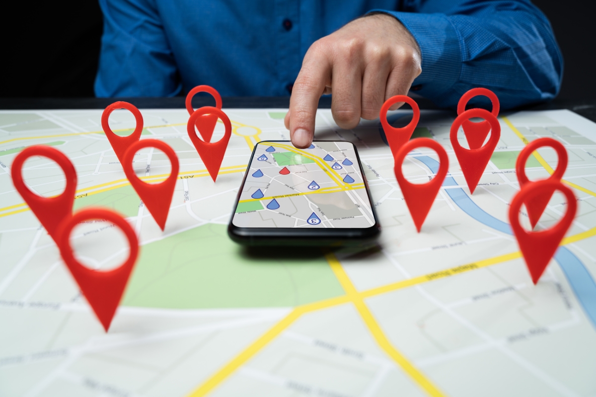 A person points at a map on a smartphone, surrounded by large red location markers on a physical map, illustrating the concept of What Is Hyperlocal Marketing.