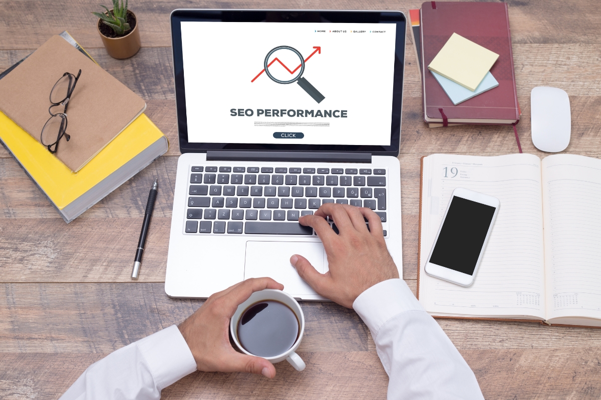 Person using a laptop showing "SEO Performance" on the screen, with a magnifying glass and graph icon. Desk has books, notepads, a smartphone, coffee cup, small plant, and a mouse. Nearby is an article titled "What Is Hyperlocal Marketing.