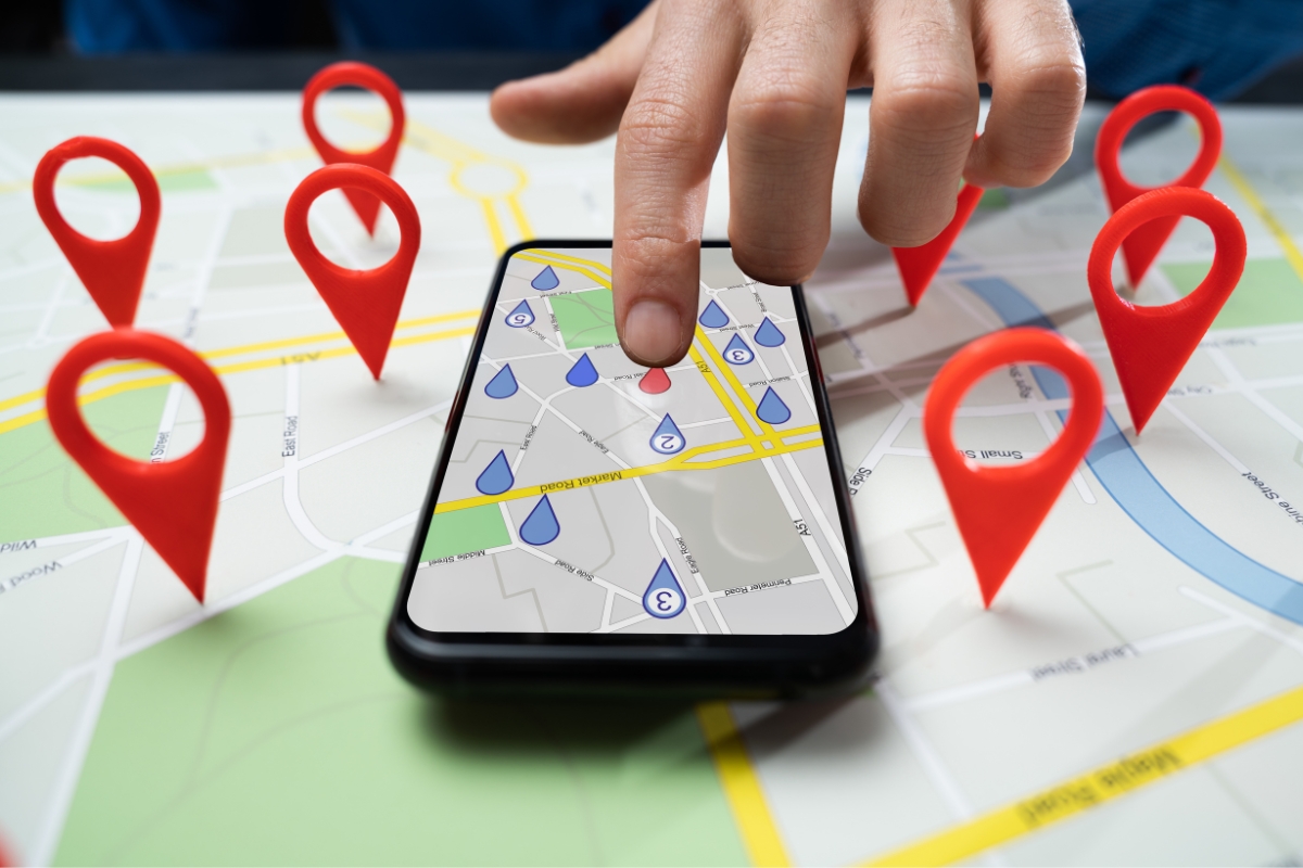 A hand points at a smartphone displaying a map with multiple location markers set on a physical map. Red location markers are also placed on the actual map around the phone, illustrating what is hyperlocal marketing in action.