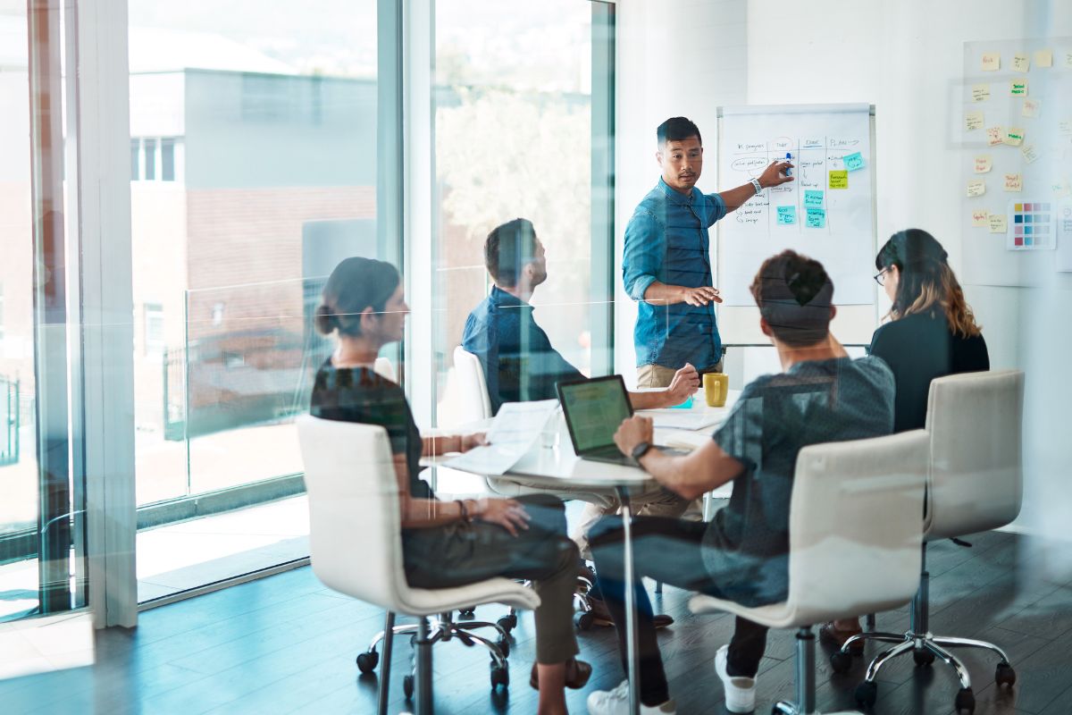 A person stands and gestures toward a whiteboard filled with notes on Inbound vs Outbound Marketing during a meeting with four seated colleagues in a conference room.