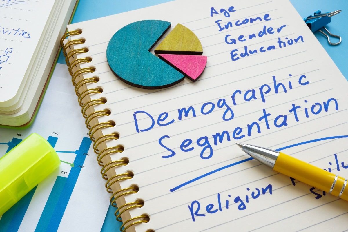 A notebook with "Demographic Segmentation" written on it, surrounded by a pie chart, a pen, a highlighter, and graphs. Age, Income, Gender, and Education are listed as factors. It's a clear nod to understanding what is market segmentation in terms of breaking down your audience by key demographics.