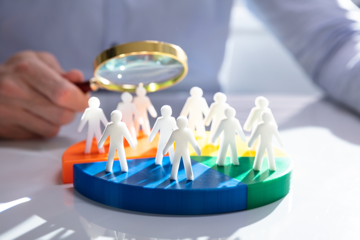 A person holds a magnifying glass over white figurines arranged on a multicolored pie chart, suggesting an examination of data or demographics to understand what is market segmentation.