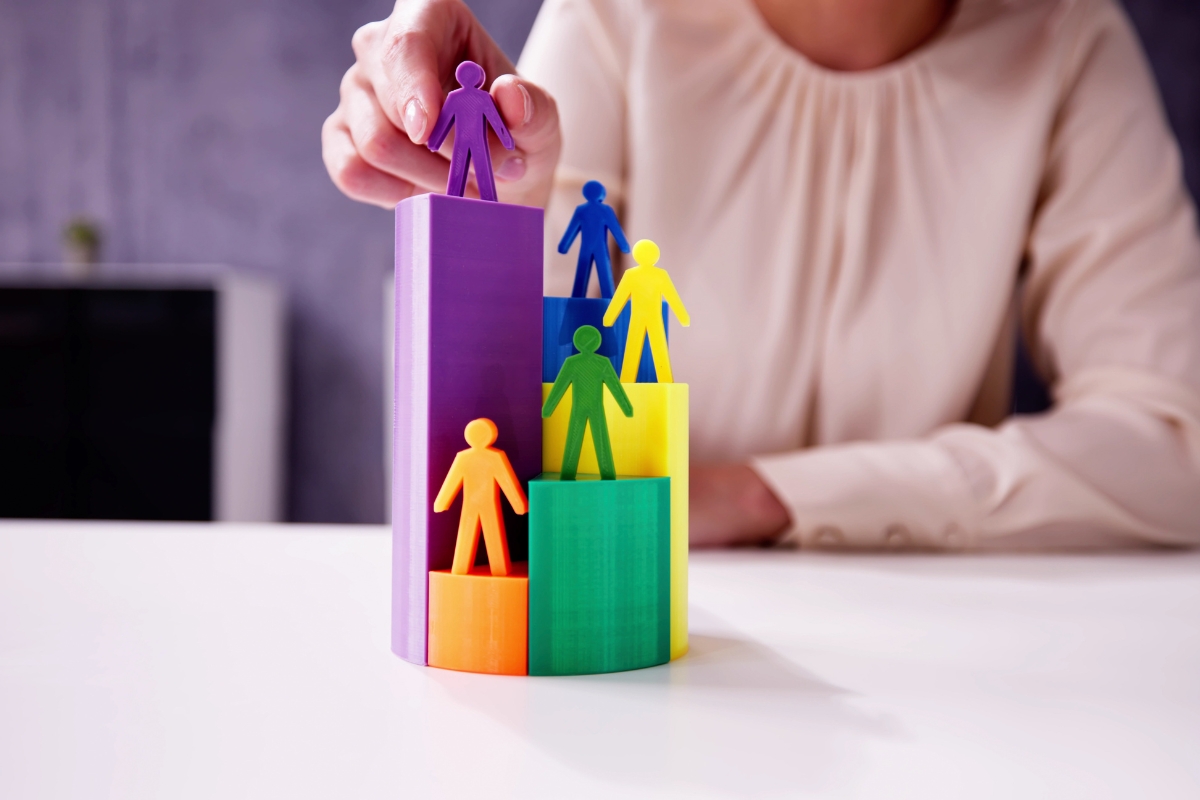 A person in a cream blouse arranges multicolored human-shaped figures on top of stacked, multicolored cylindrical blocks, a visual representation often used in marketing to explain what is market segmentation.