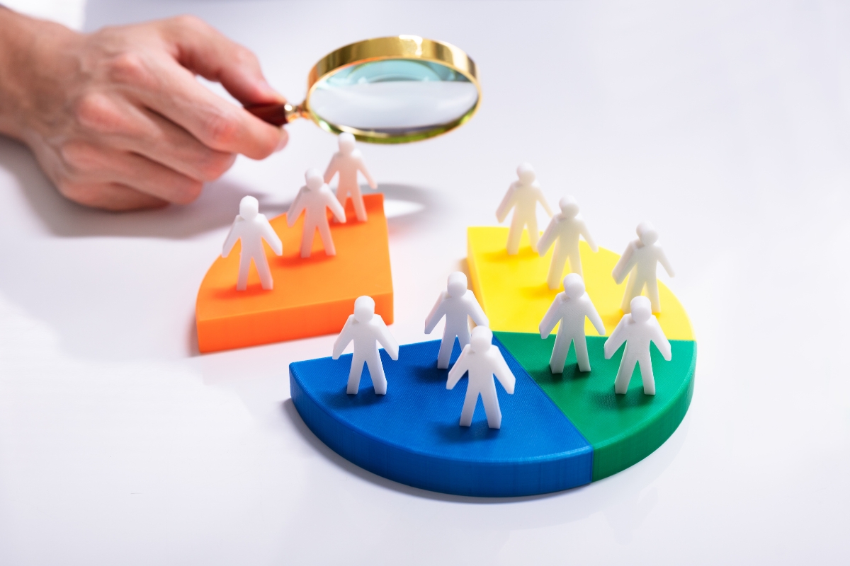 A hand holding a magnifying glass over plastic human figures on colorful pie chart segments: orange, yellow, blue, and green illustrates what is market segmentation.