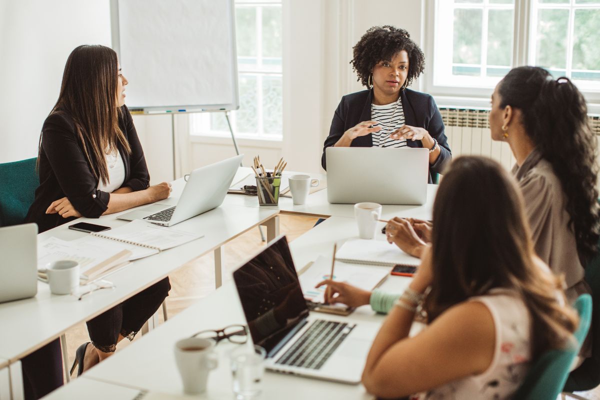 Four women are sitting around a table in an office setting, engaging in a discussion on inbound vs outbound marketing. Laptops, notebooks, and coffee cups are scattered on the table, with a whiteboard serving as the backdrop.