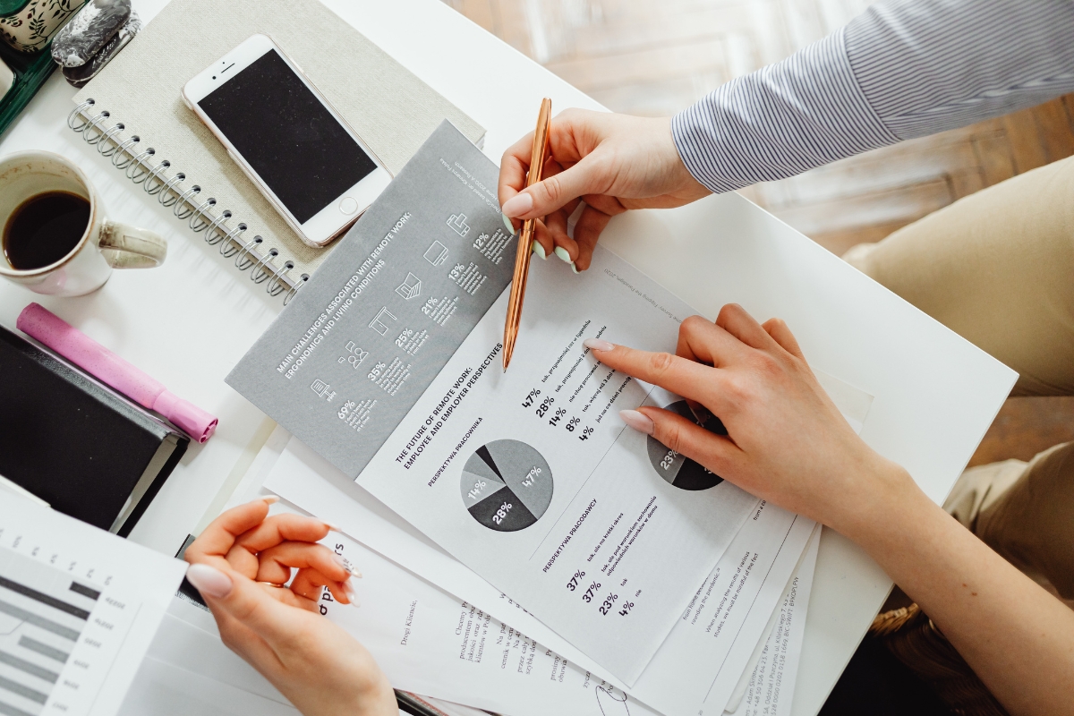 Two people are discussing printed charts and graphs at a desk cluttered with office supplies, a smartphone, and a coffee cup. As they delve into the data, they touch on strategies and principles of what is relationship marketing, emphasizing the importance of customer connections.