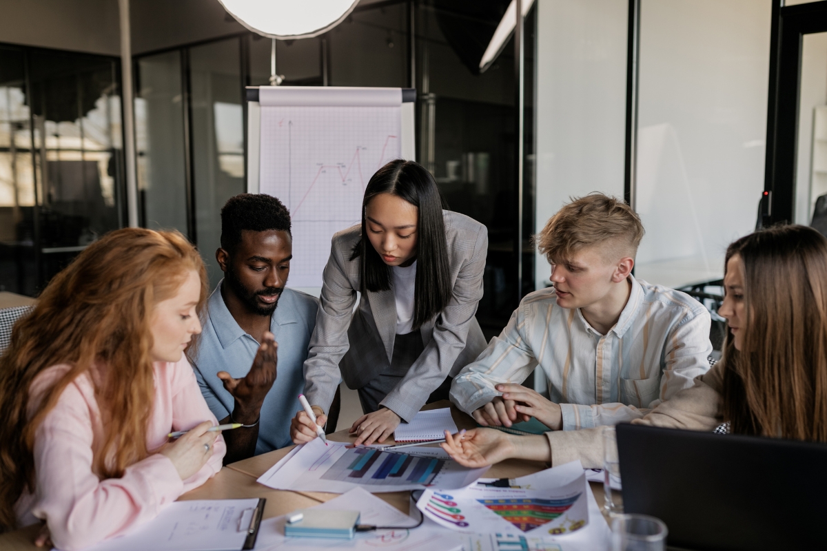 A group of five people are gathered around a table covered with charts and papers in an office setting, actively discussing and examining documents on what is relationship marketing.