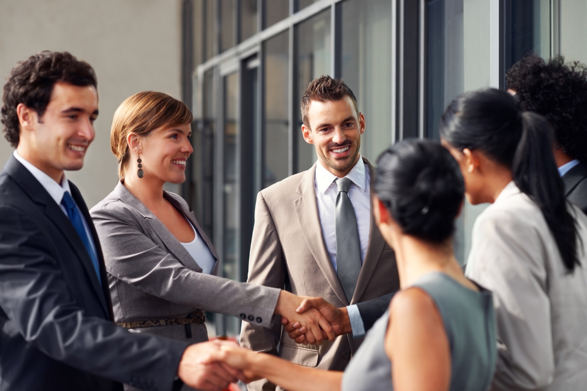 A group of business professionals in formal attire are standing outdoors, shaking hands, and smiling during a friendly interaction—exemplifying what is relationship marketing. They are positioned near a glass building.