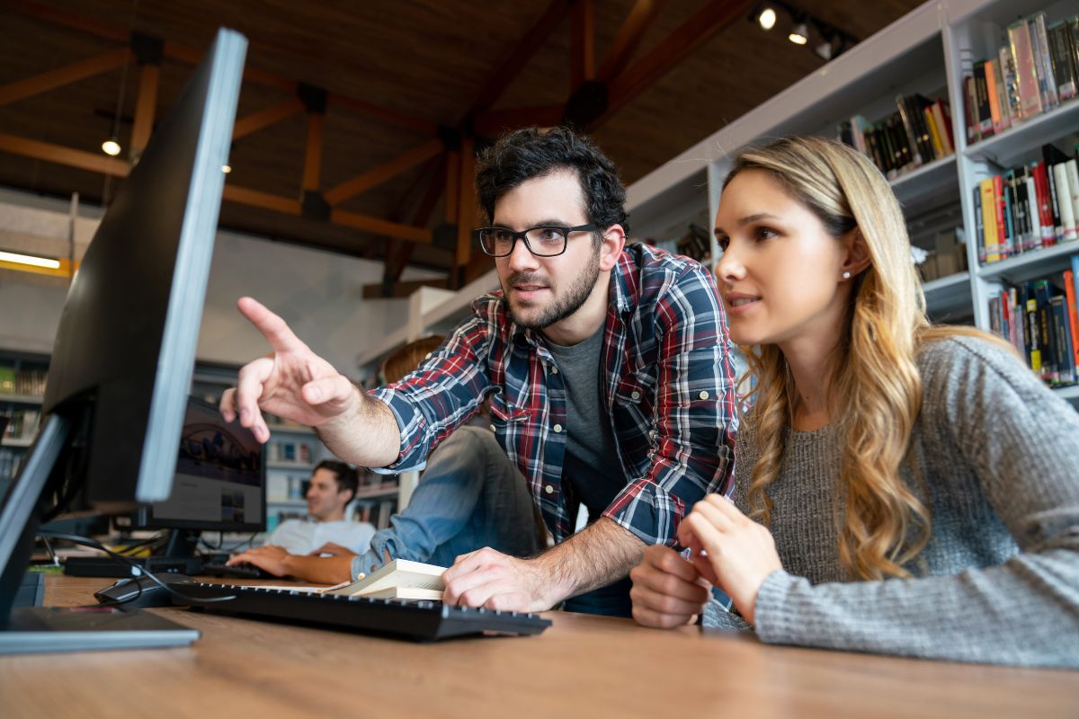 Two people look at a computer screen in a library, with one pointing at the monitor and discussing "What Is Semantic Search." Shelves of books are visible in the background.