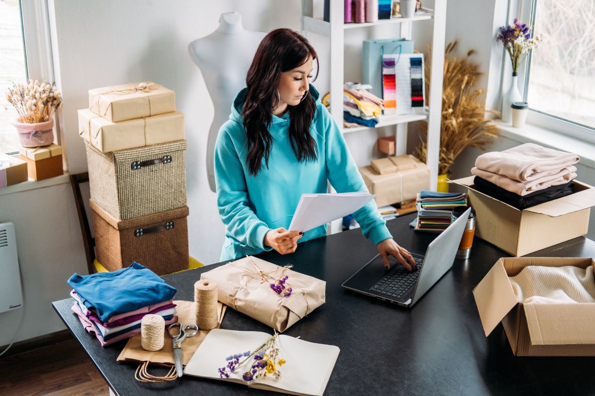 A person in a teal hoodie working on a laptop, possibly researching what is SEO, surrounded by gift boxes, wrapping materials, folded clothes, and dried flowers on a table in a well-lit room with shelves in the background.