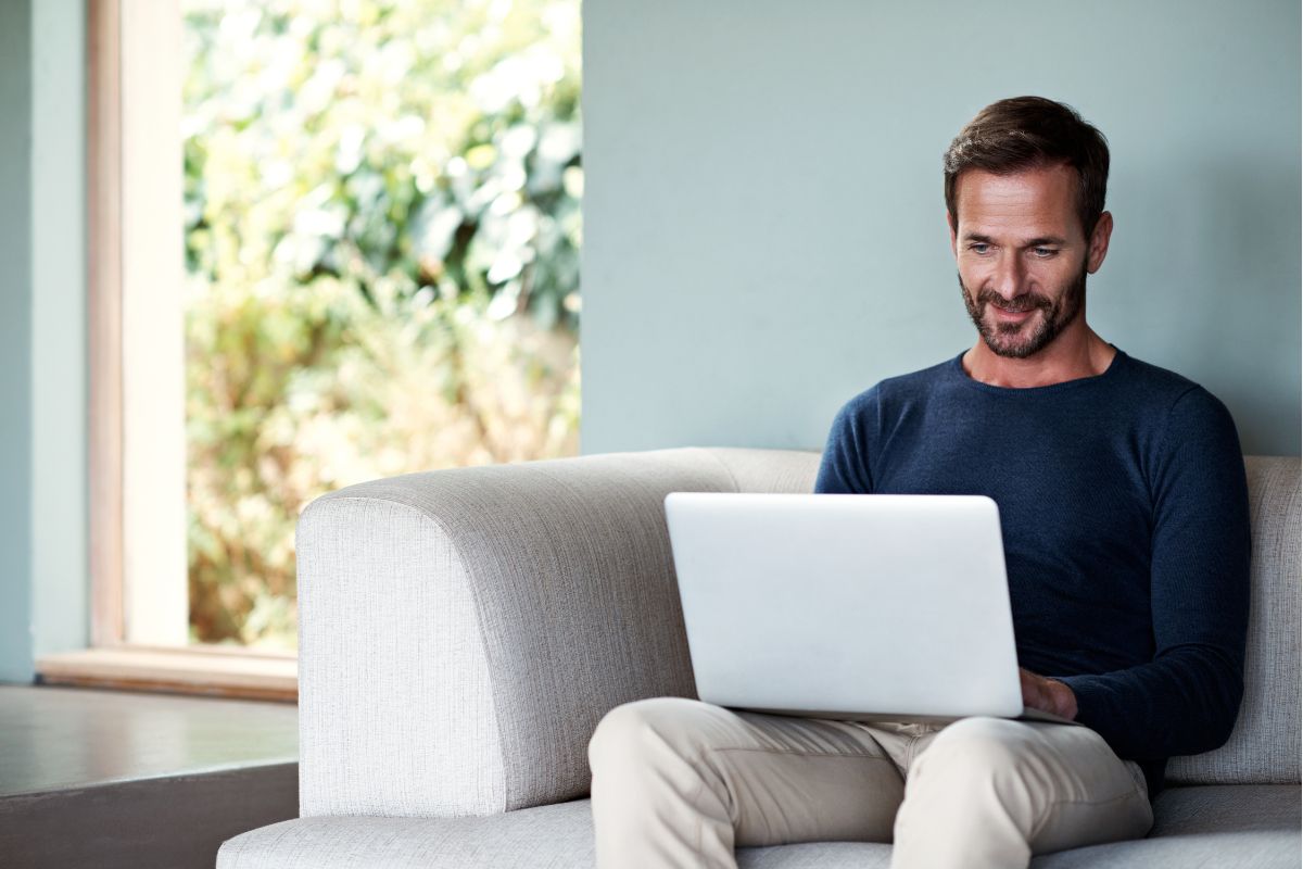 A man sits on a beige couch, working on his laptop with a smile on his face. He wears a dark long-sleeve shirt and khaki pants, perhaps researching how to create an email signature. A large window with greenery outside is in the background.