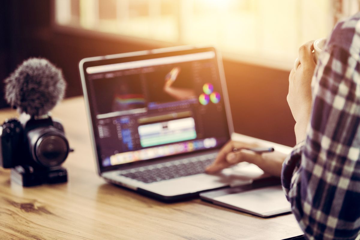 A person is working on video editing software on a laptop, crafting content about how to use YouTube Shorts for business. Nearby, a camera and microphone rest on the wooden table, ready to capture engaging clips for YouTube Shorts.