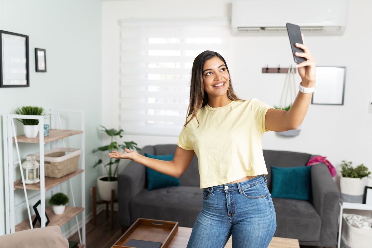 A woman wearing a yellow shirt and jeans takes a selfie in a living room adorned with plants, a gray sofa, wall art, and her favorite "How to Use Youtube Shorts for Business" guide on the coffee table.