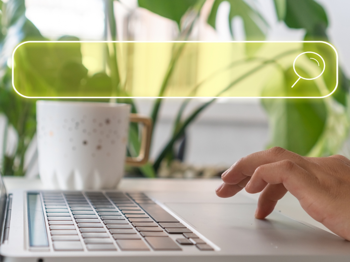 A hand typing on a laptop keyboard with a white coffee mug in the background. A translucent search bar with a magnifying glass icon overlays the image, hinting at keyword search and optimization techniques. Green plants are visible in the background.