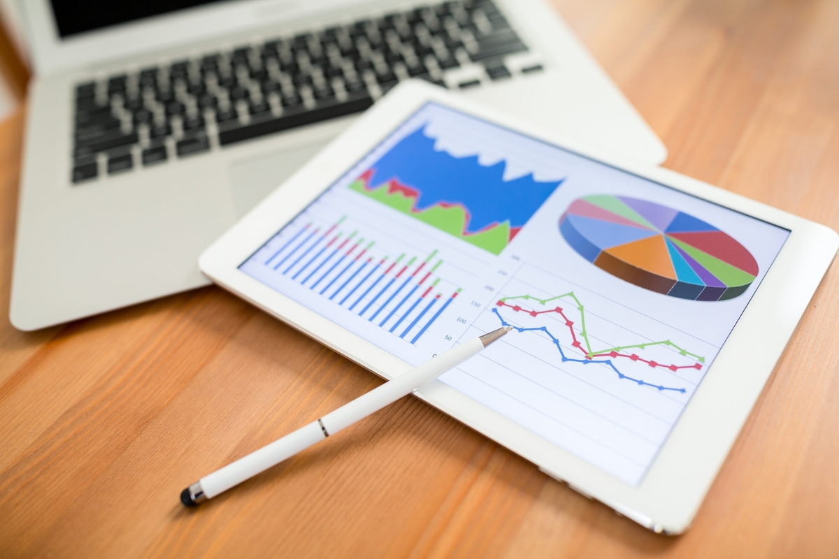 A tablet displaying colorful graphs and charts related to AI in marketing analytics rests on a wooden table, with a stylus on top. A laptop is in the background.