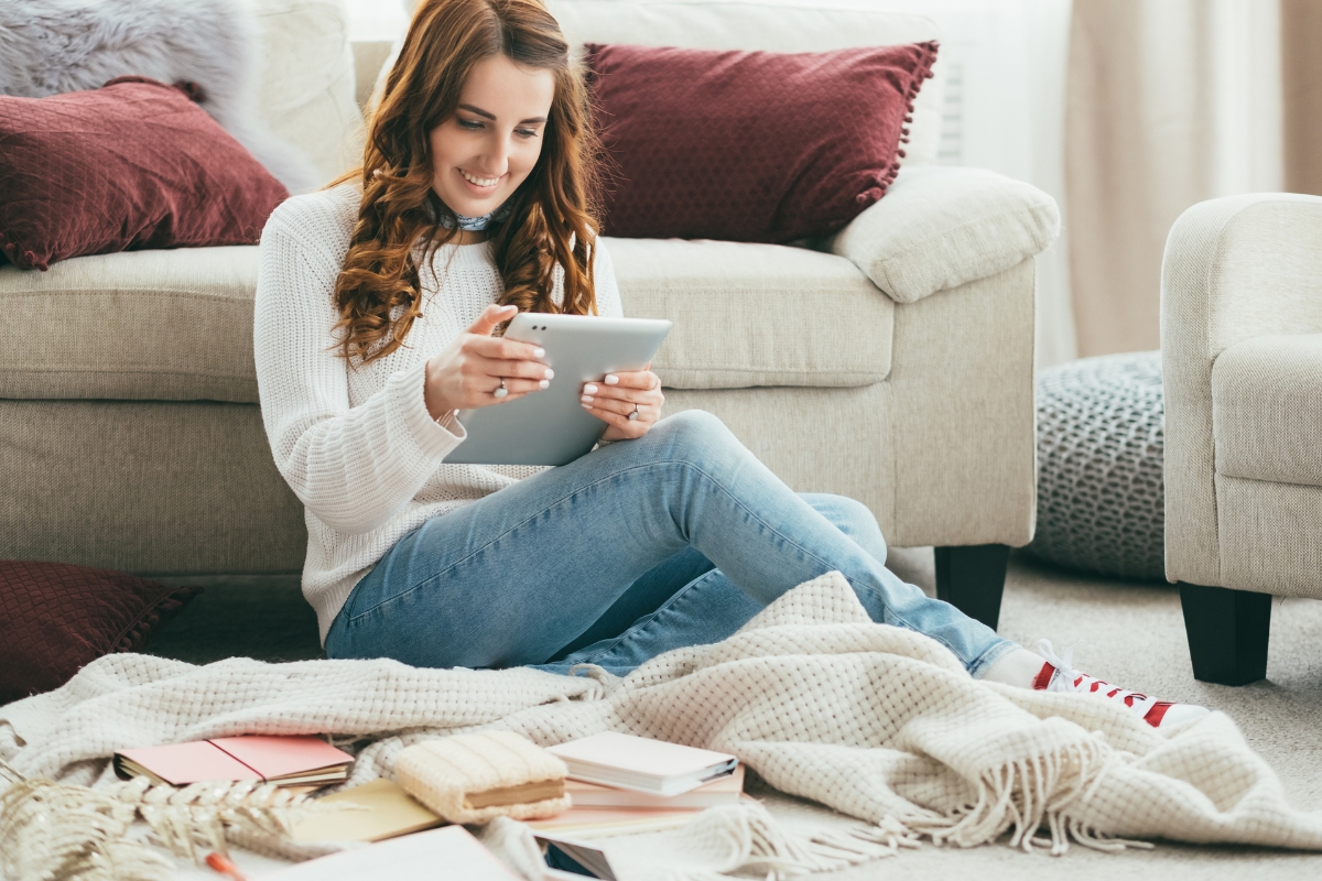 A woman sits on a blanket on the floor, smiling while using a tablet. She is surrounded by books and notebooks, delving into ai in marketing analytics, with a cozy couch and pillows in the background.