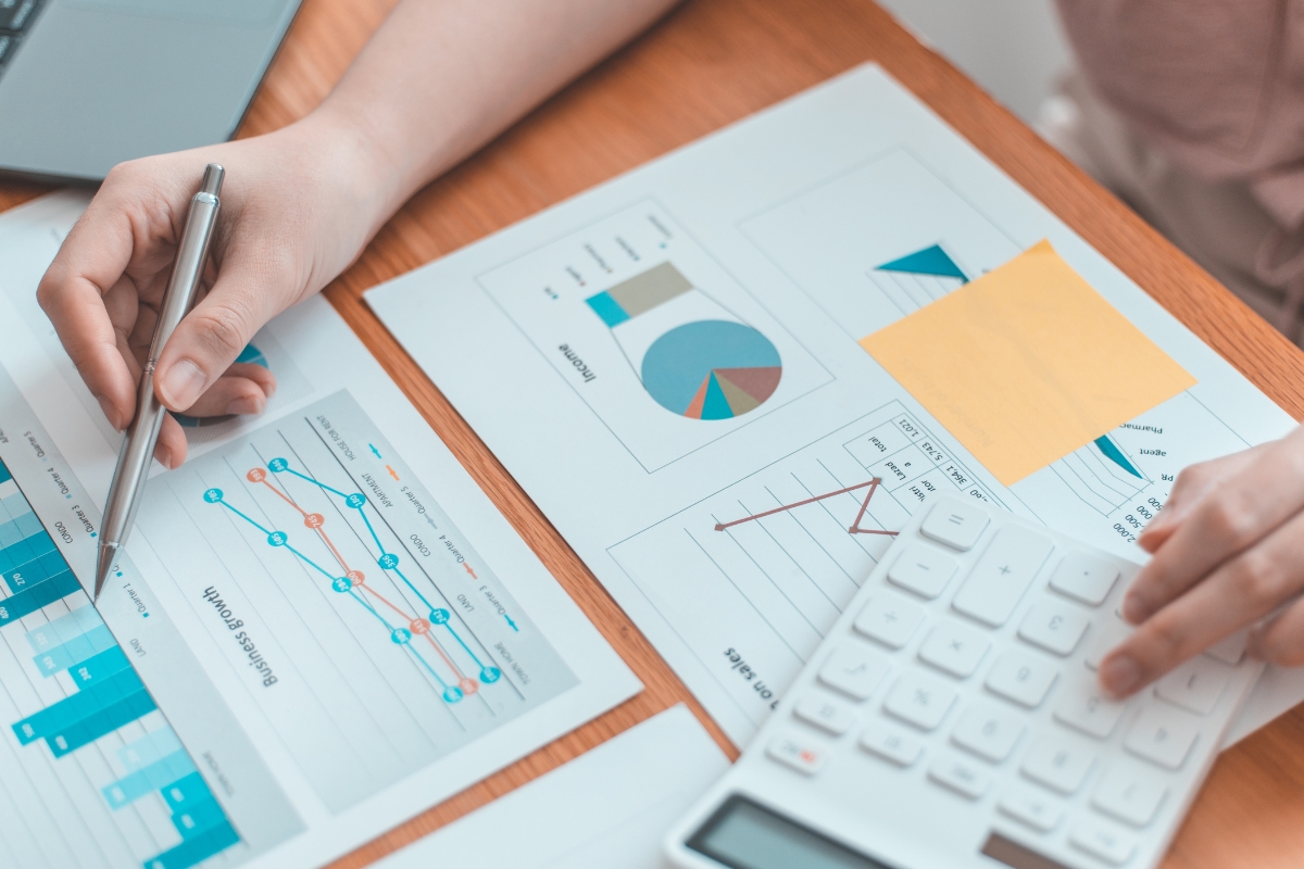 A person is analyzing graphs and charts on paper, using a calculator while writing notes on a desk, seamlessly integrating AI in marketing analytics to enhance data insights.