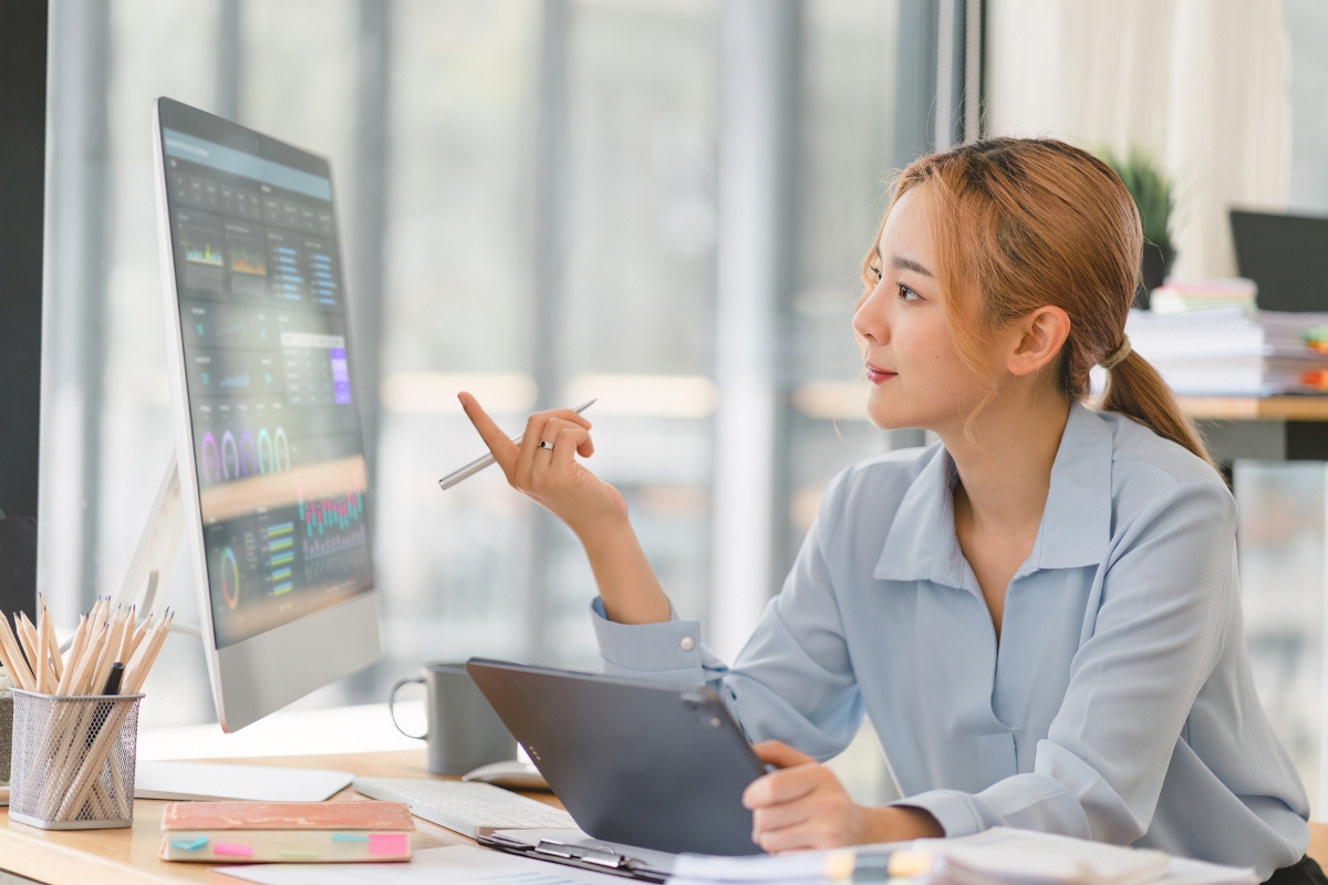 A person sits at a desk, using a computer while holding a pen and a tablet. The computer screen displays various charts and graphs powered by AI in marketing analytics. The workspace is organized with office supplies and books.