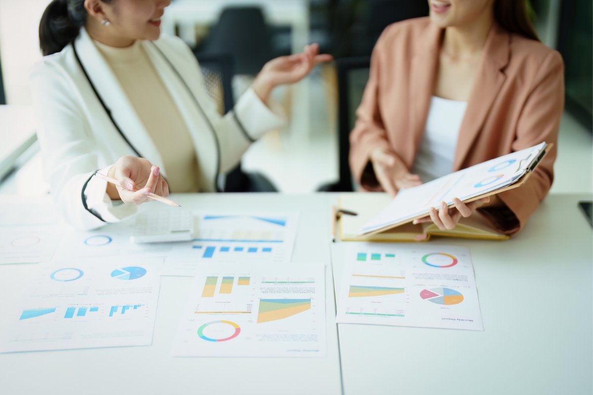 Two women in a meeting review charts and graphs with one holding a notebook and the other gesturing. Various printed charts, illustrating the impact of AI in marketing analytics, are spread out on the table.