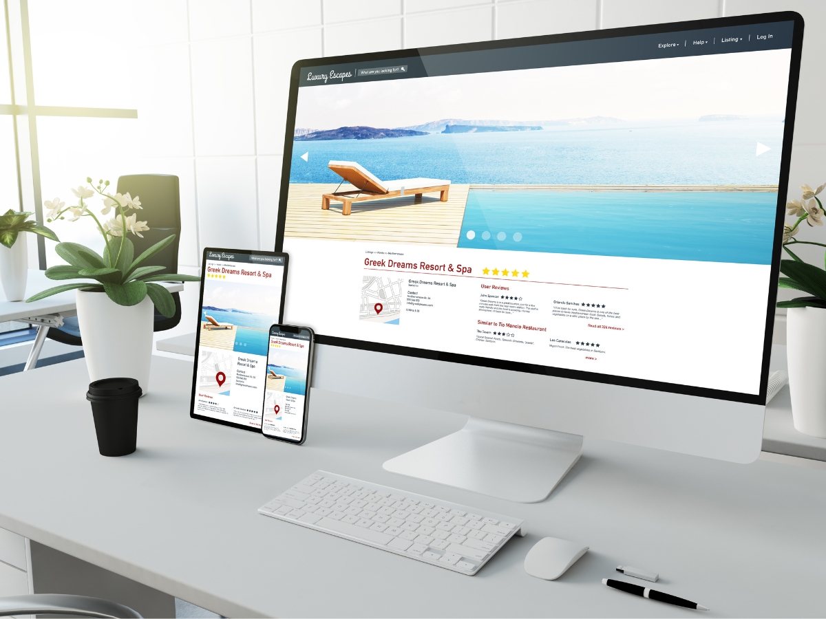 Desktop computer, smartphone, and tablet displaying a travel website featuring a beach resort. The desk hosts a keyboard, mouse, coffee cup, office supplies, and materials for citation building. A large window serves as the backdrop.