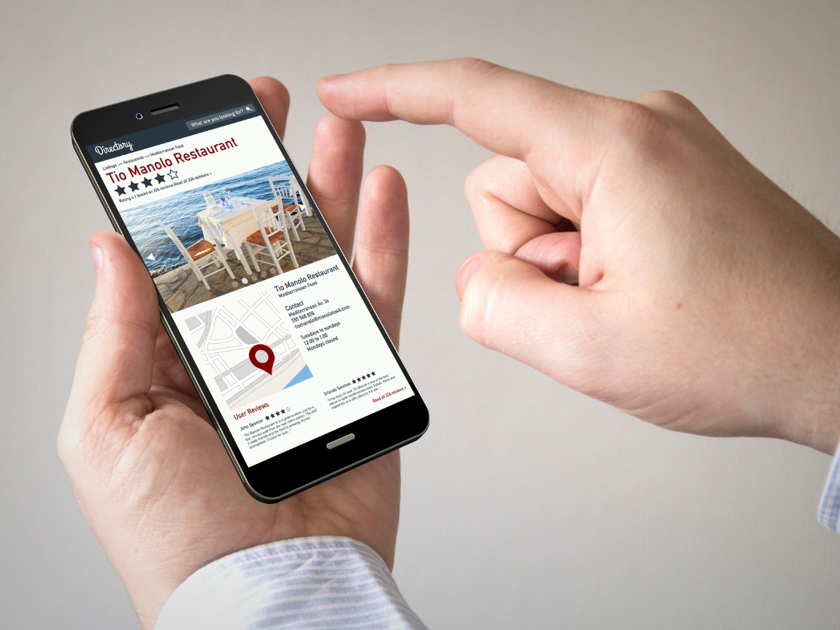 A person holding a smartphone and pointing at an online restaurant listing for "Tio Manolo Restaurant," featuring an image of outdoor seating and a map location, highlights the importance of citation building for local SEO.