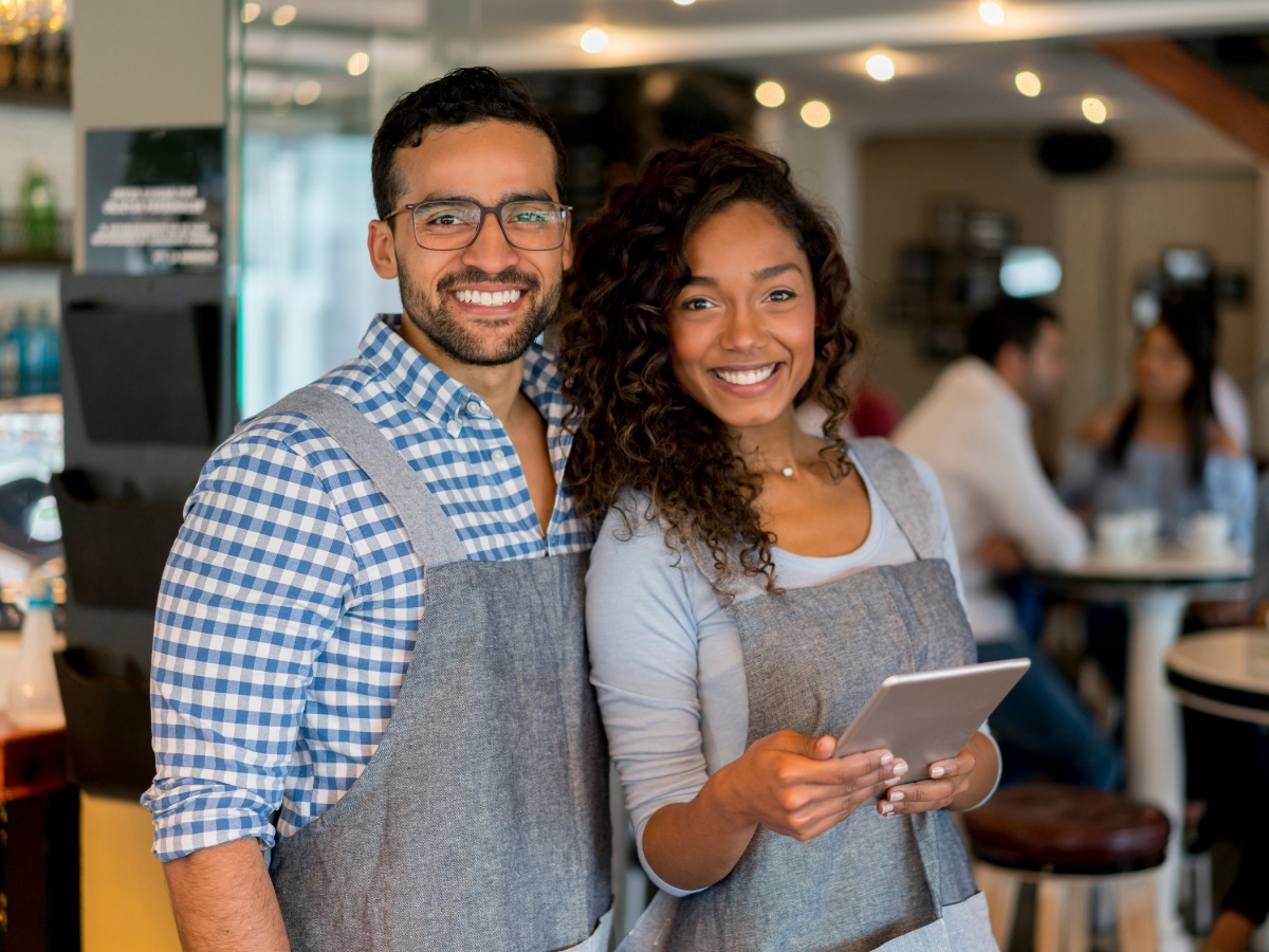 Two smiling individuals, one holding a tablet, are wearing aprons and standing in what appears to be a busy café or restaurant, possibly discussing citation building for their online menu.