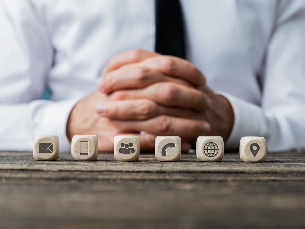 A person with hands folded sits behind a table with six wooden blocks displaying icons: envelope, smartphone, group of people, phone, globe, and location pin — tools essential for effective citation building.