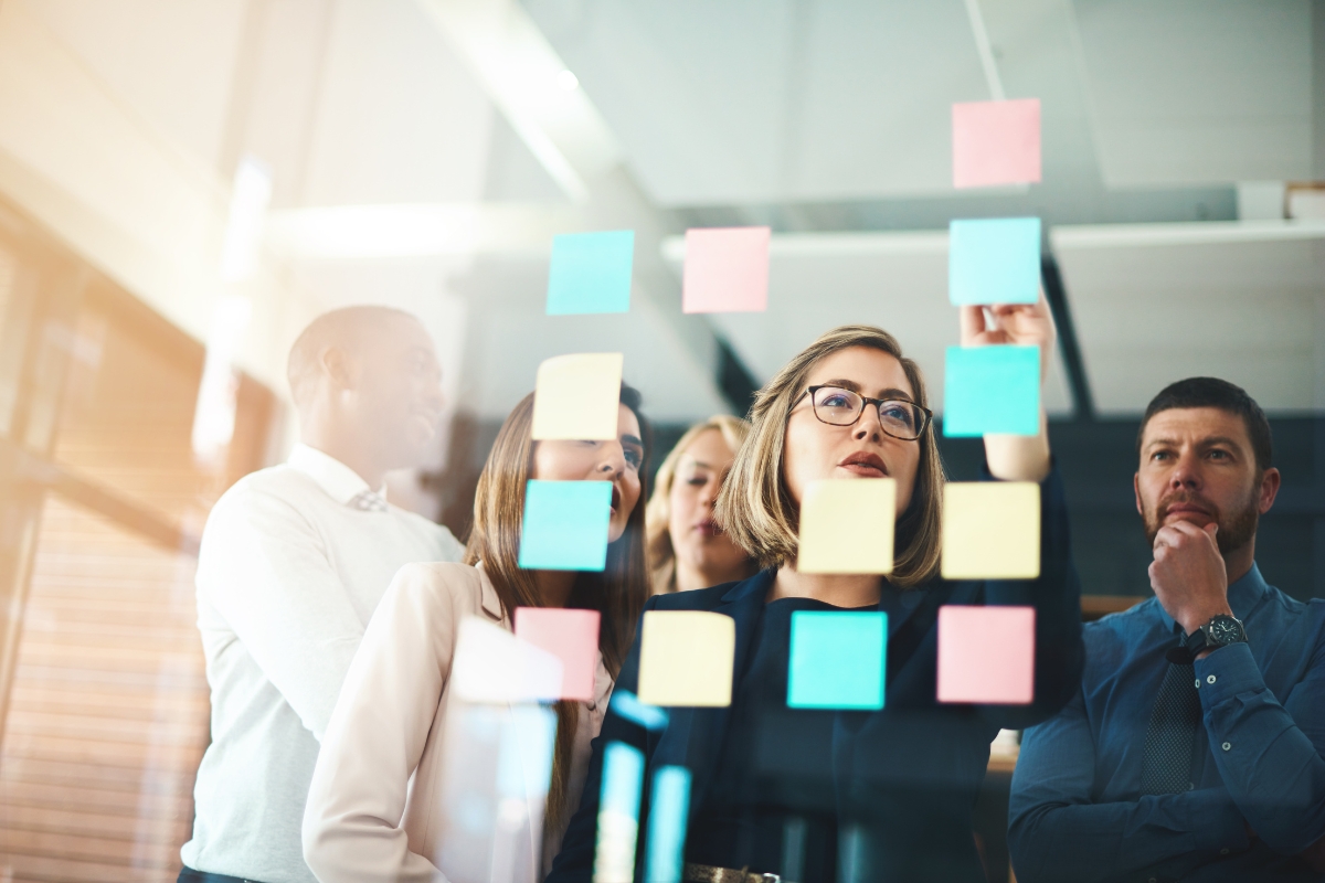 A group of people stand behind a glass wall covered with colorful sticky notes, discussing how to come up with content ideas. One person is pointing at a note while others attentively listen and think.