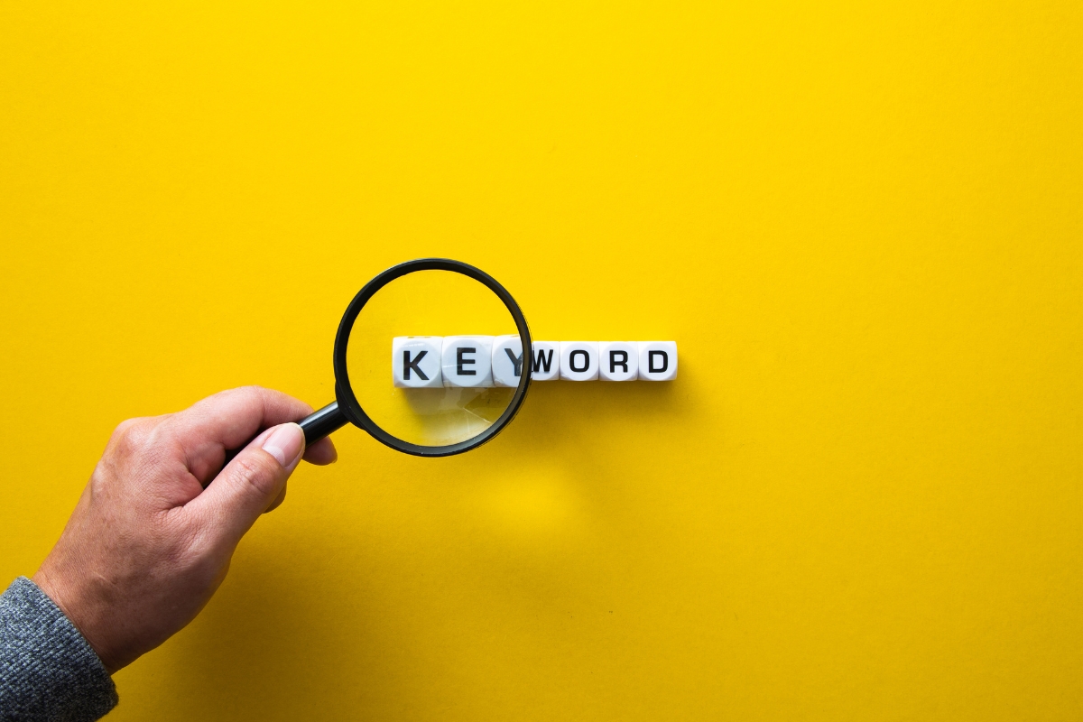 Hand holding magnifying glass over the word "KEYWORD" made with white letter tiles on a yellow background, illustrating how to come up with content ideas.