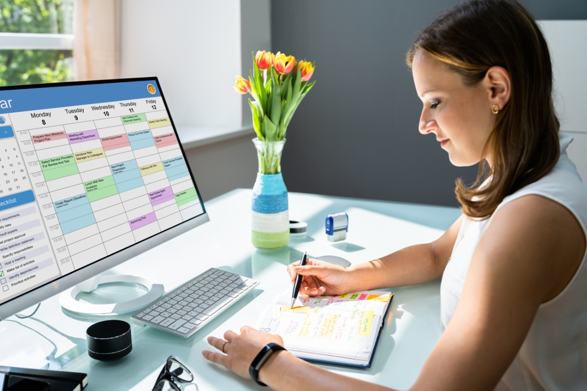 A woman is sitting at a desk, working on a planner with a pen. A large computer screen displaying a color-coded calendar, likely filled with tips on how to come up with content ideas, is in front of her. A vase of tulips brightens the desk.