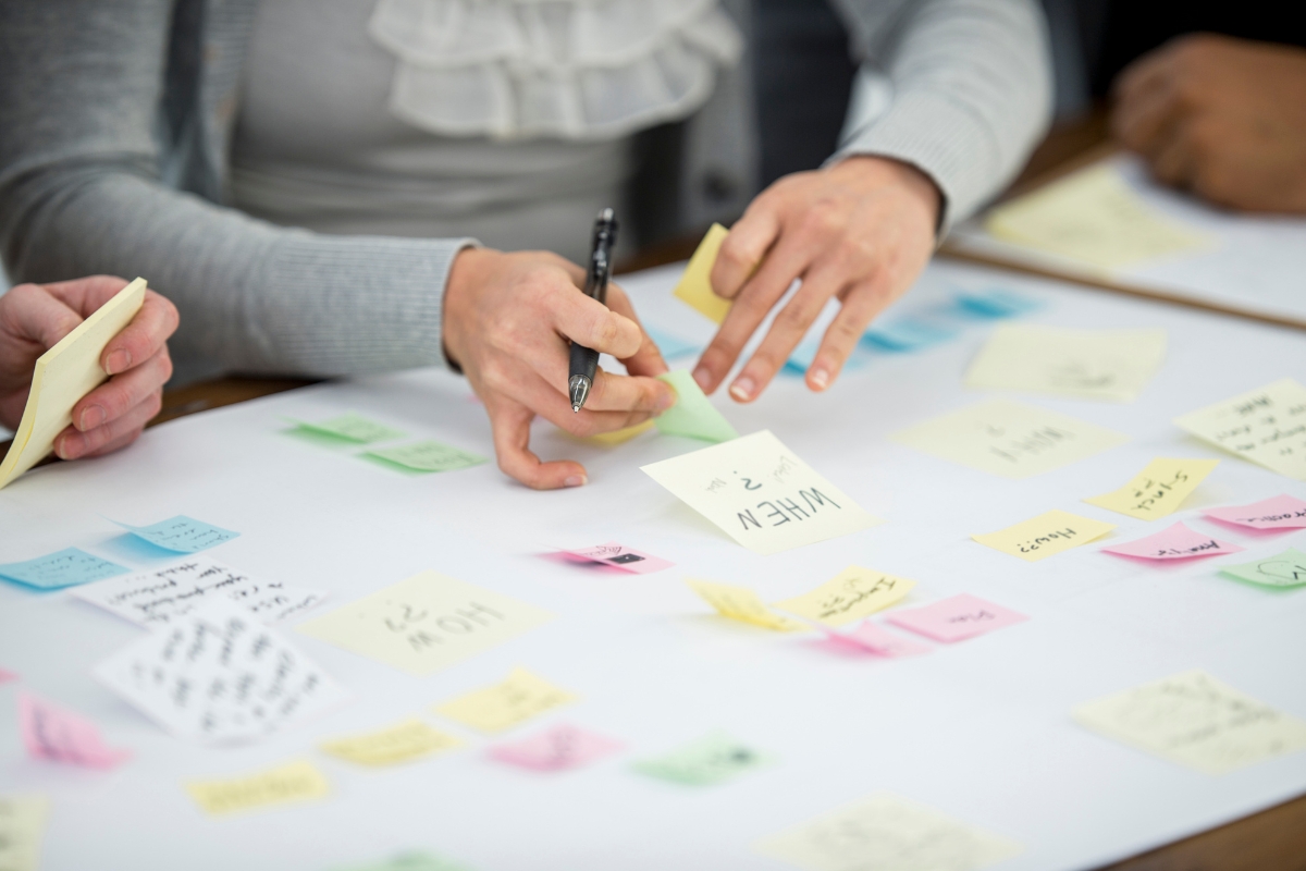 Several people collaborate on a project, using sticky notes and a large paper on a table, brainstorming how to come up with content ideas. One person writes on a yellow sticky note with a pen.