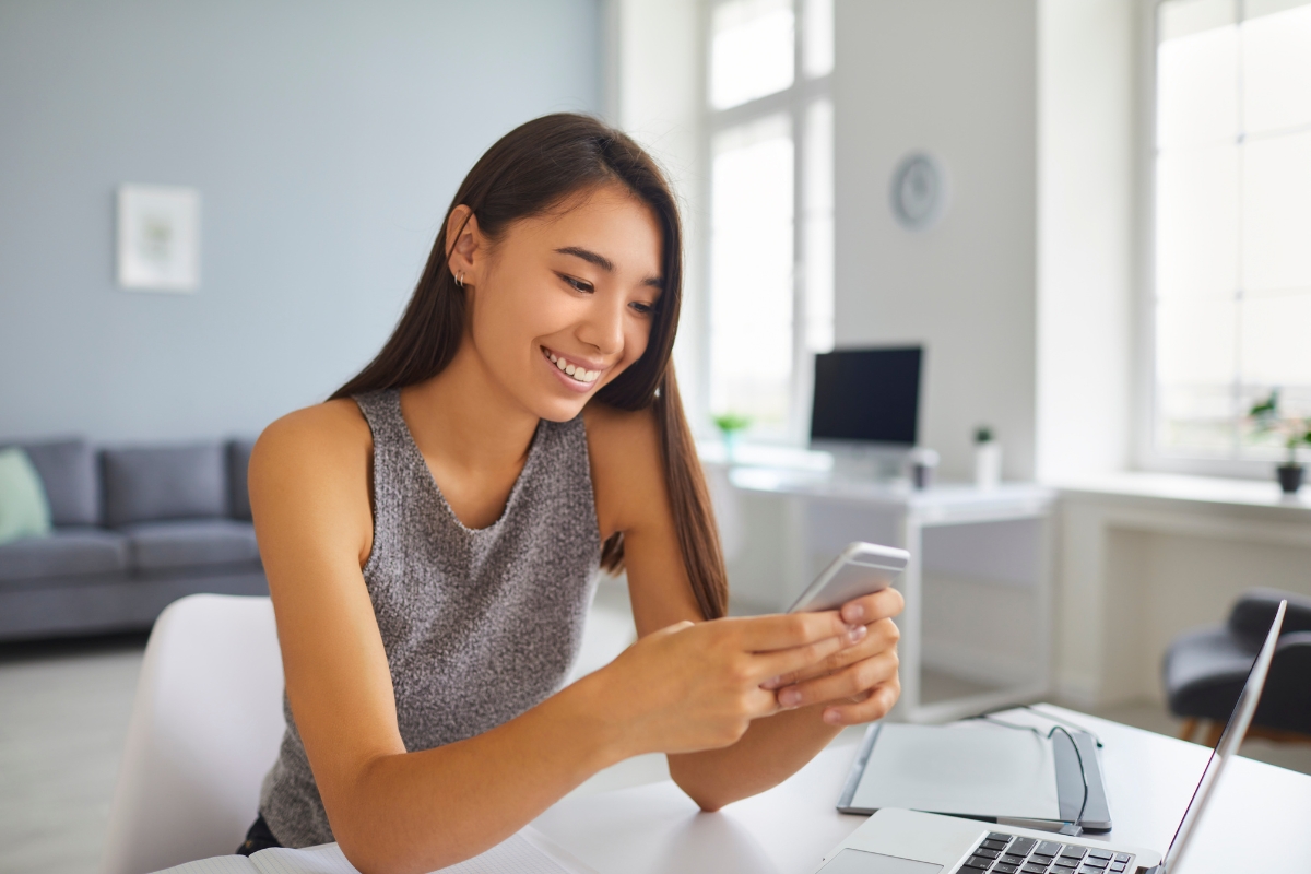 A woman is smiling and using her smartphone while sitting at a table with a laptop and papers, possibly researching how to create buyer personas, in a modern, well-lit room.