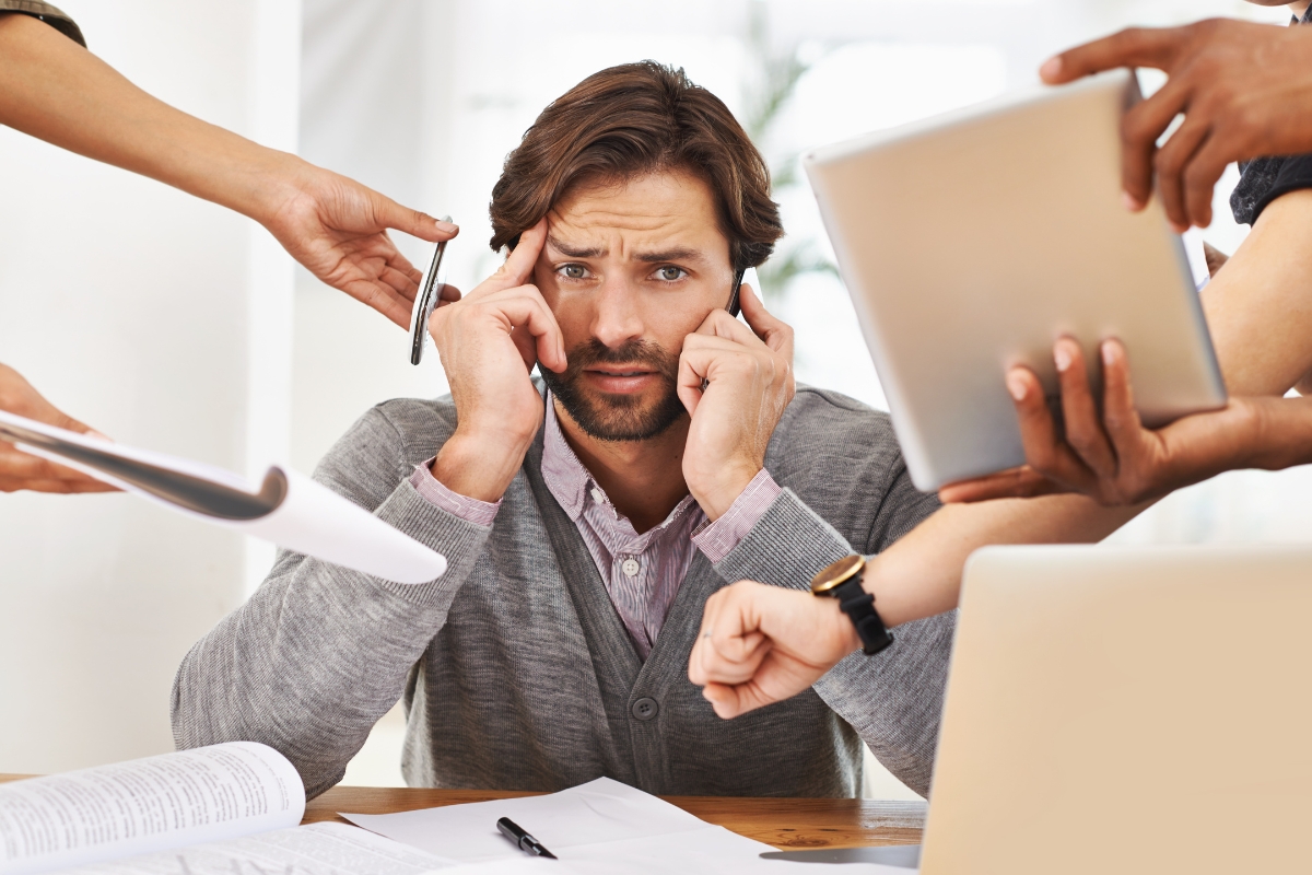 A stressed man sits at a desk, surrounded by people handing him papers on how to create buyer personas, a tablet, and a pen, while he holds his temples and talks on the phone.