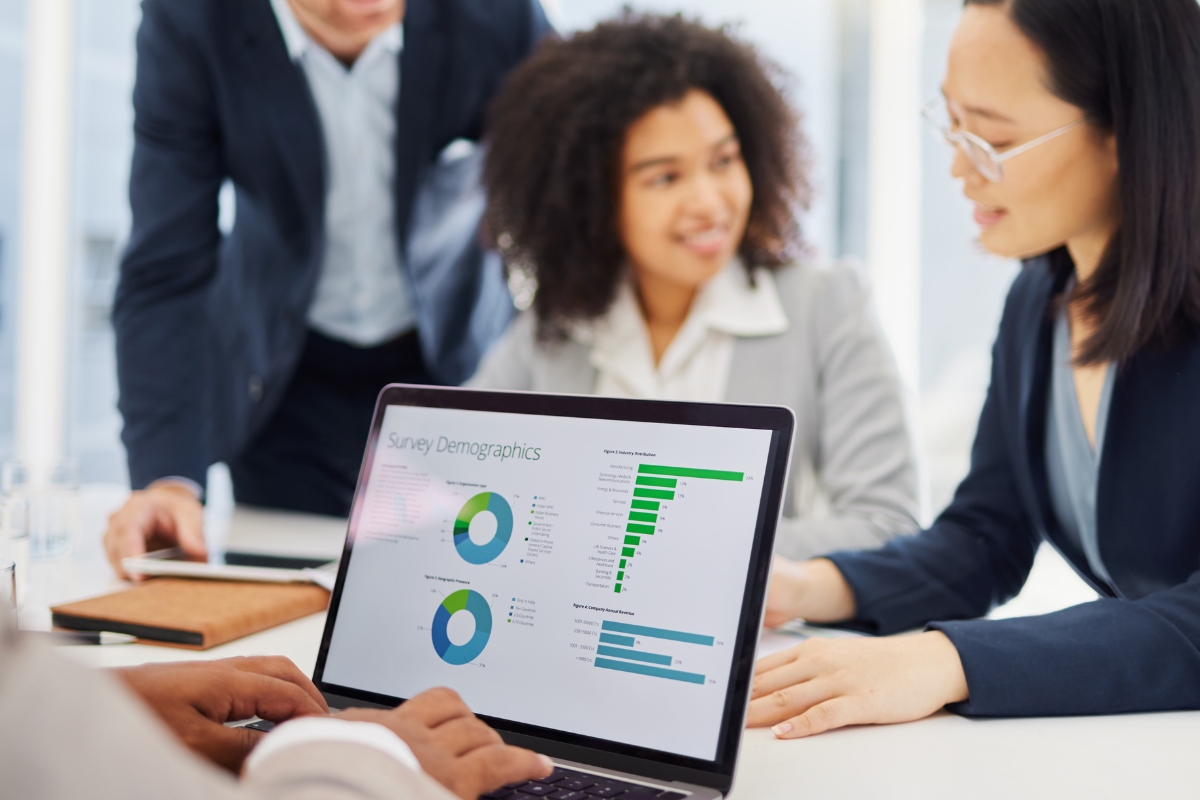 A laptop with survey demographics charts is on a desk, with three people in business attire discussing how to create buyer personas in the background.