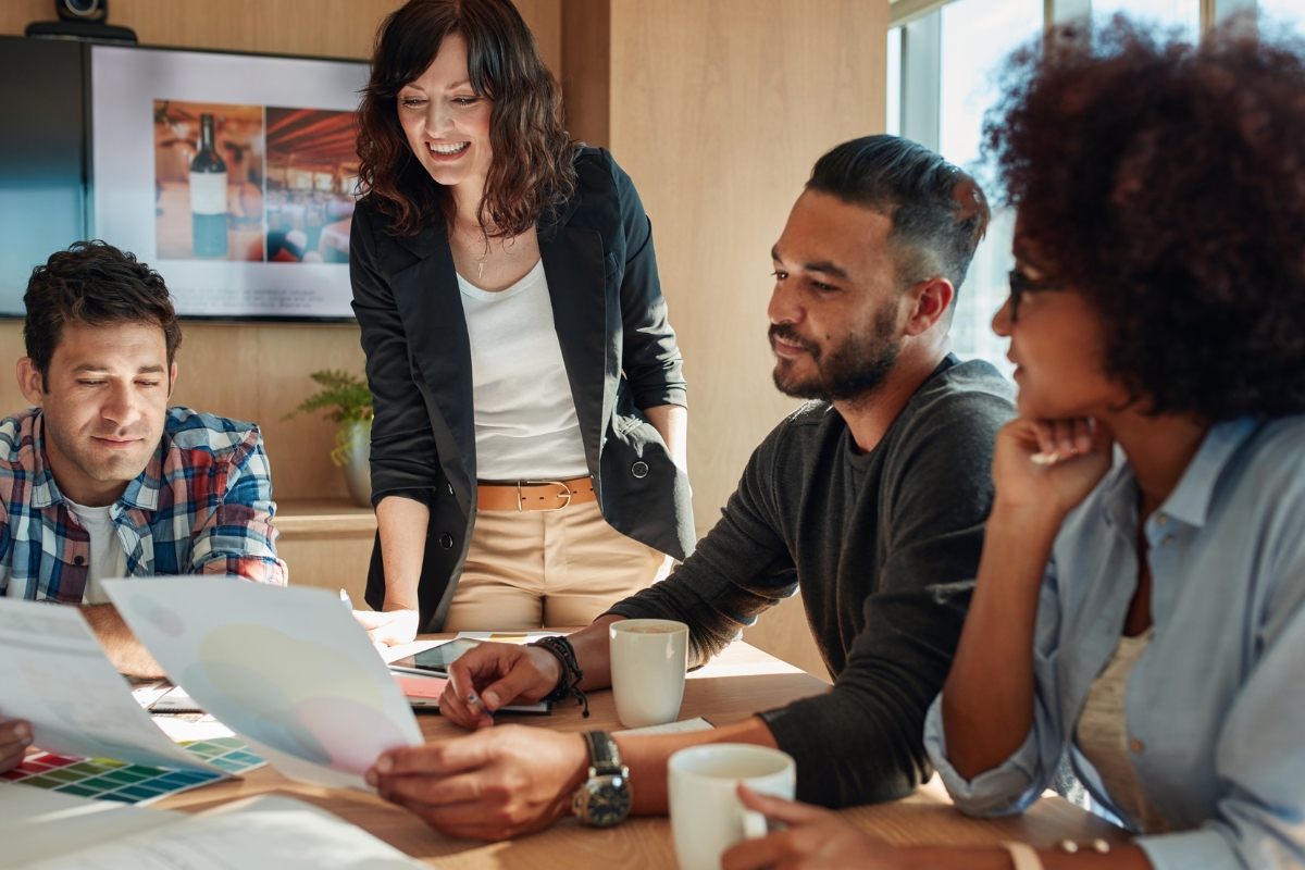 Four people in a meeting room reviewing documents on how to create buyer personas. One person is standing and three are seated, all holding papers or cups. A framed picture and a window are in the background.