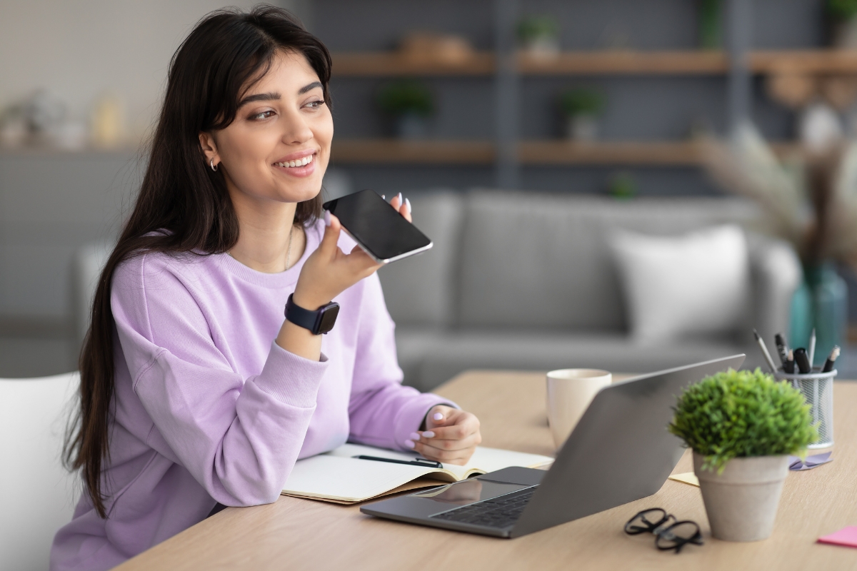 A woman in a purple sweater holds a smartphone and speaks into it, possibly researching how to get featured snippets, while sitting at a desk with a laptop, notebook, and cup. Shelves and a sofa are visible in the background.