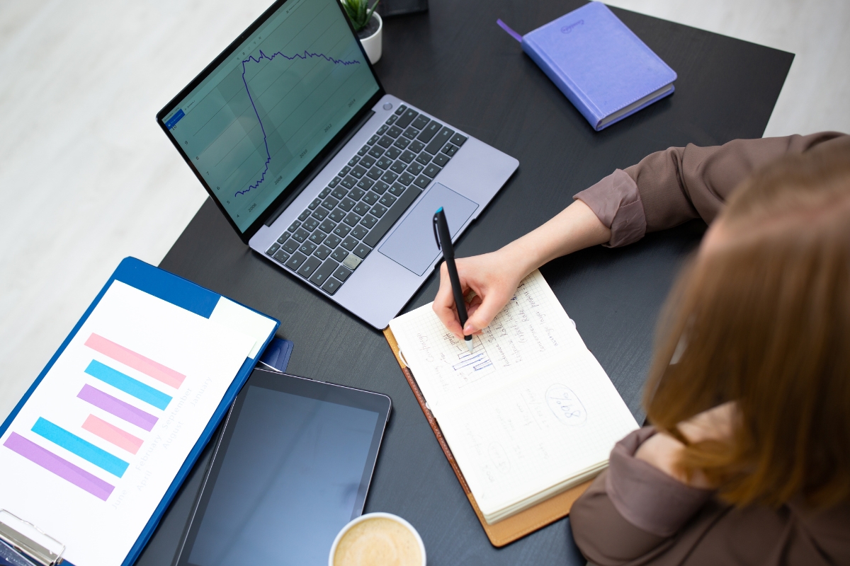 Person taking notes from charts displayed on a laptop and documents, with a tablet, coffee, a purple notebook, and research on how to get featured snippets on the desk.